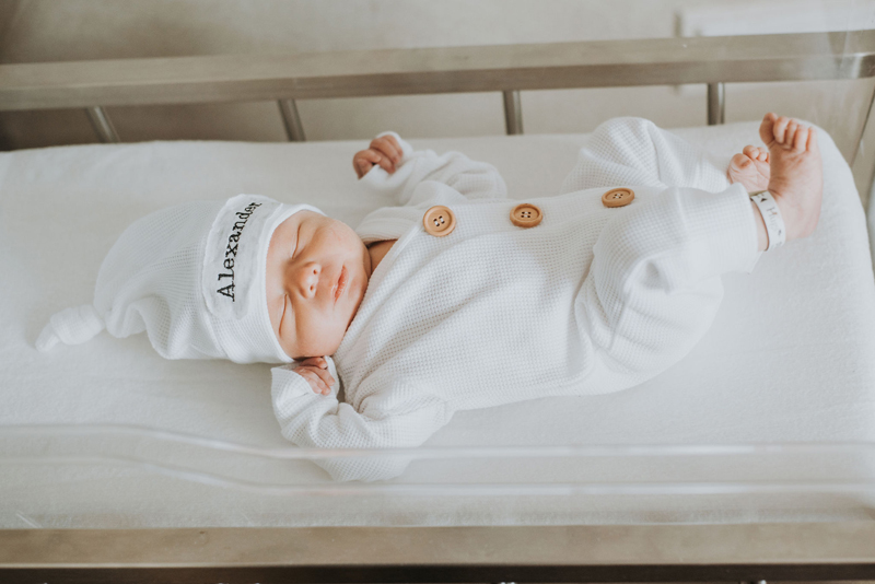 Newborn Photographer, a baby lays in a hospital crib with a white onesie and a white beanie that reads "Alexander"