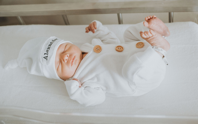 Newborn Photographer, a baby lays on clean sheets in a small crib with a beanie that reads "Alexander"