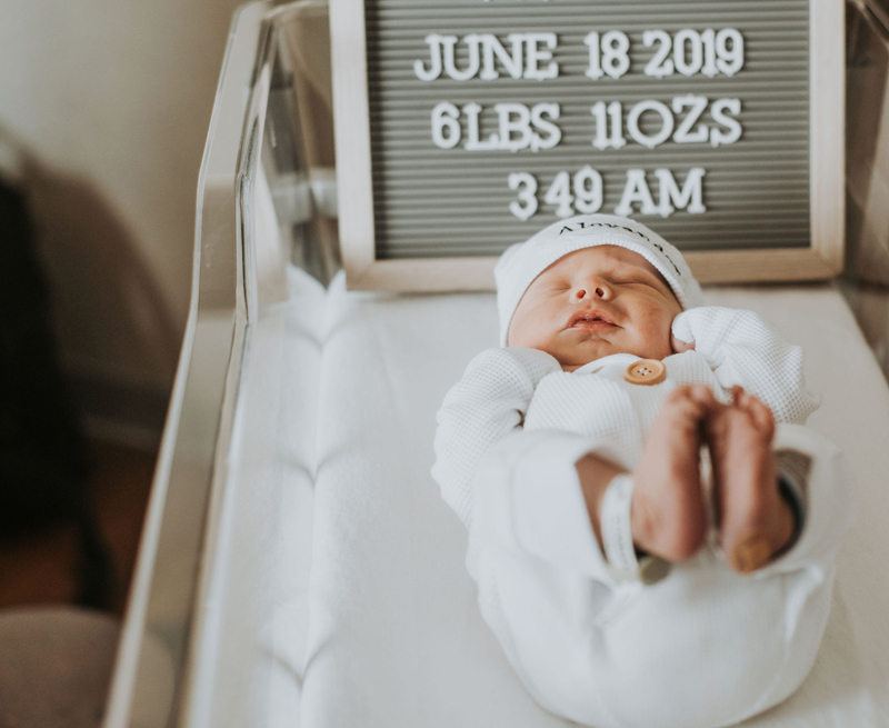 Newborn Photographer, a baby lays in a hospital bassinet with small felt board behind him that reads "June 18 2019, 6lbs 11oz, 3:49am"