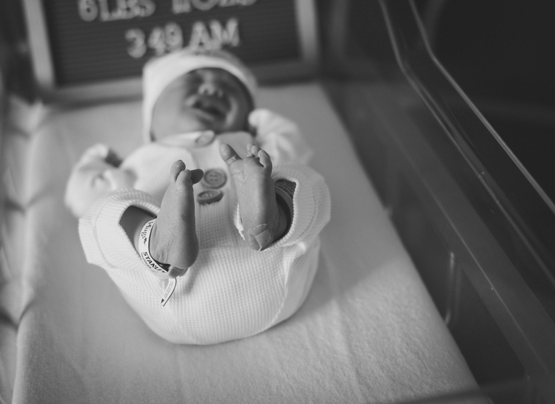 Newborn Photographer, a baby lays on a recovery room scale, legs in the air, crying, wearing a warm white onesie