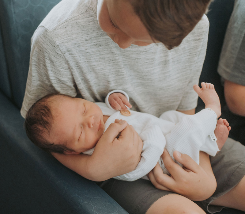 Newborn Photographer, an older sibling holds his newborn baby sibling on the couch
