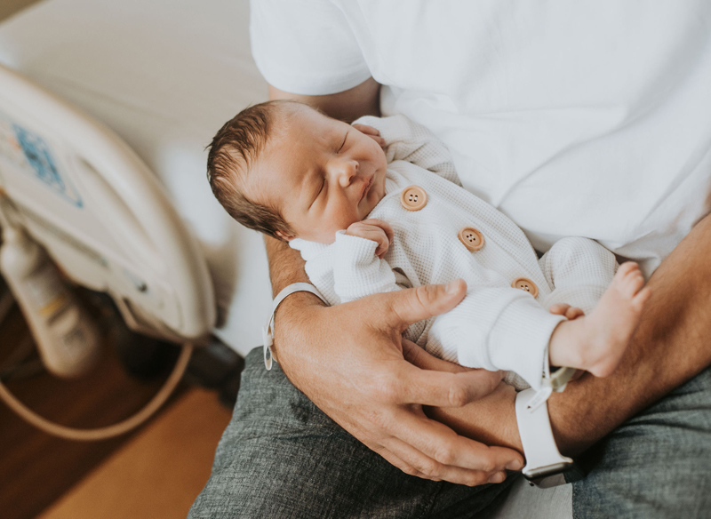 Newborn Photographer, a fathers arms hold his newborn baby in his lap as he sleeps comfy and secure