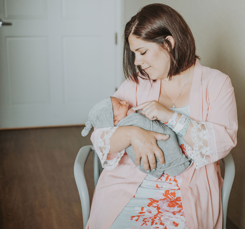 Newborn Photographer, a mother sits in a chair holding her baby and touching his little fingers