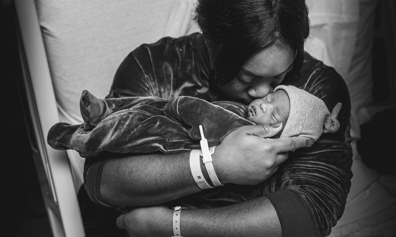 Newborn Photographer, mom gives her newborn baby a tender squeeze in the hospital bed
