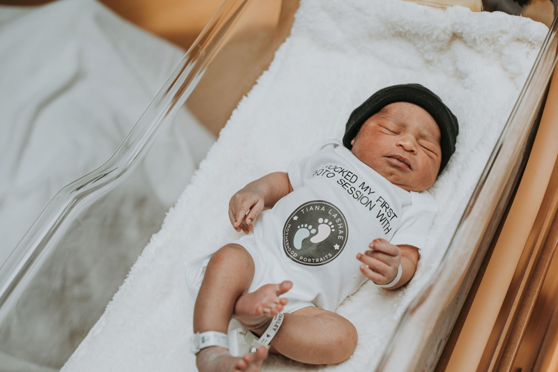 Newborn Photographer, a baby lays sleeping cozily in a hospital cradle with a onesie that reads "Booked my first Photo Session with Tiana Lashae"
