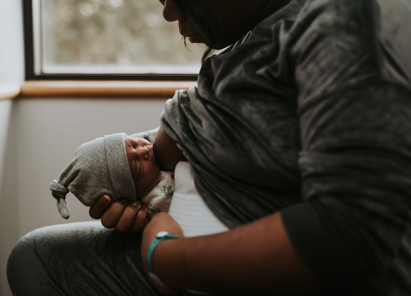Newborn Photographer, a mother breastfeeds her baby in the hospital recovery room, baby wears a warm beanie and is wrapped in a hospital baby blanket