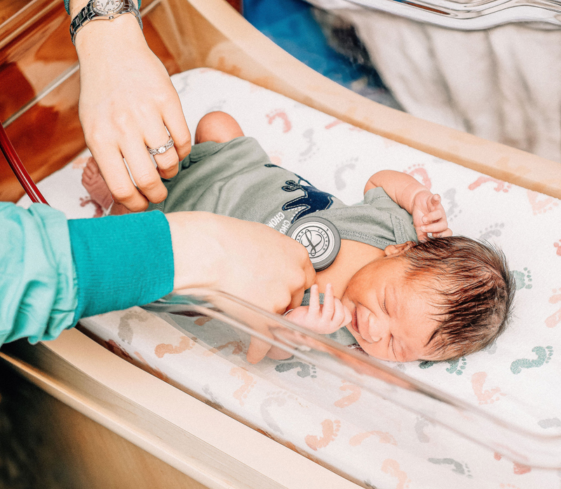 Newborn Photographer, a little baby boy lays sleeping in a hospital crib as family reaches into to say hello