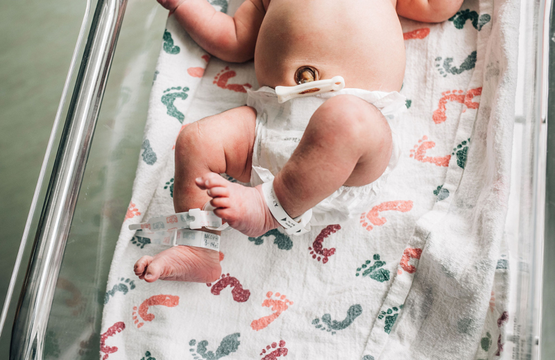 Newborn Photographer, a baby lays in hospital cradle on a patterned sheet with footprints, hospital bracelets on the baby's ankles