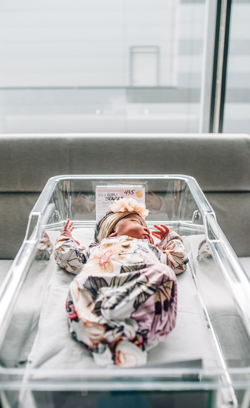 Newborn Photographer, a small baby lays sleeping in a hospital bassinet with a colorful floral onesie