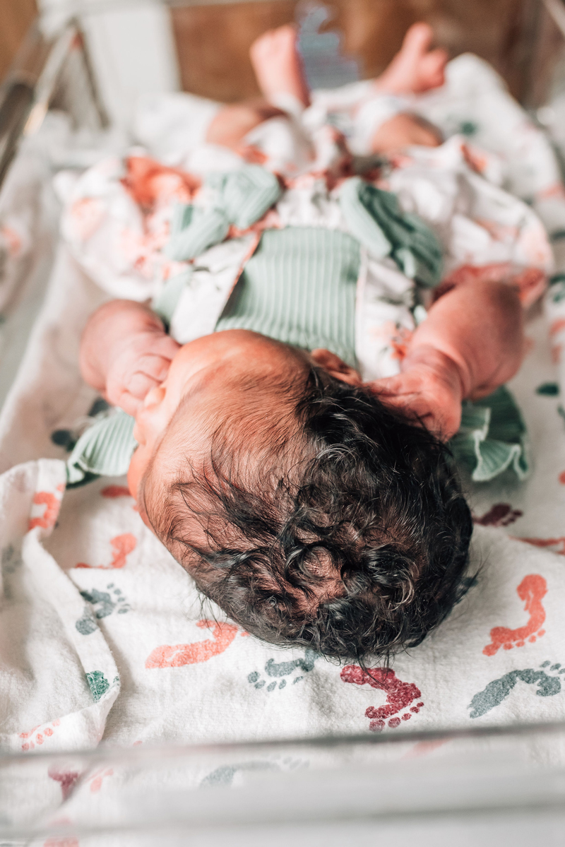 Newborn Photographer, a baby lays sleeping in a hospital bassinet with a full curly head of hair