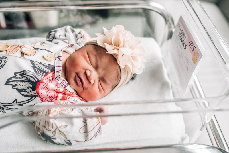 Newborn Photographer, a little baby girl sleeps in a hospital cradle and scale, she has a floral onesie and headband