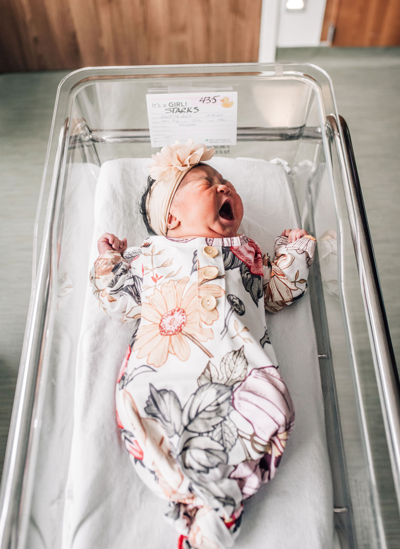 Newborn Photographer, a little baby girl cries in a hospital bassinet, she wears a floral onesie and bow headband on her head